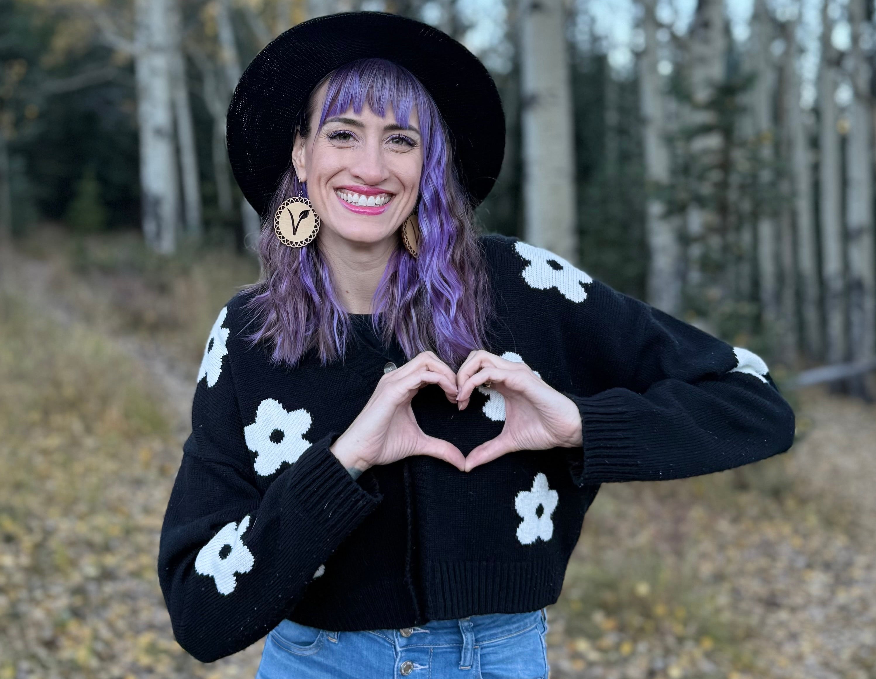 Blake Freedom  wearing a black rim hat and black sweater with white floral patterns, gesturing heart hands, standing outdoors with trees in the background.